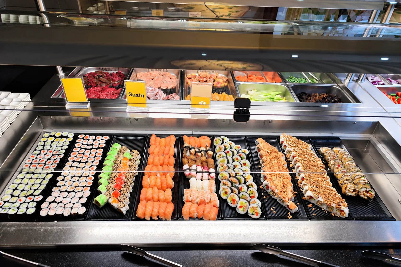 Sushi counter at the buffet: rows of maki, inside-out rolls and salmon nigiri on black trays, a yellow “Sushi” sign.