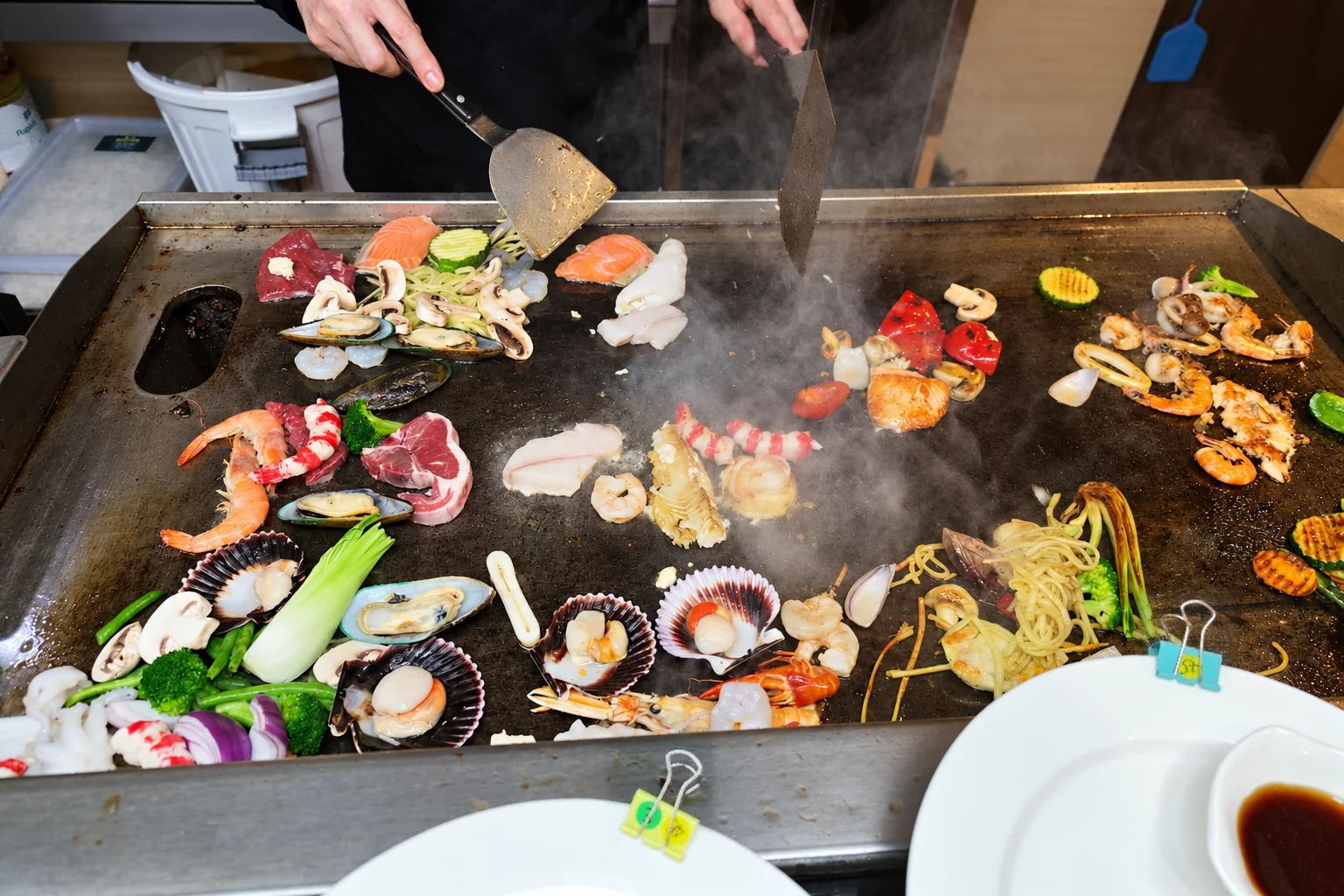 Prawns, scallops, mussels, mushrooms and vegetables on a steaming metal plate, two hands with spatulas, white plates in front.